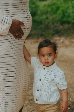 Small child wearing a white crocheted cardigan stands next to someone in a matching white dress by a sandy path.