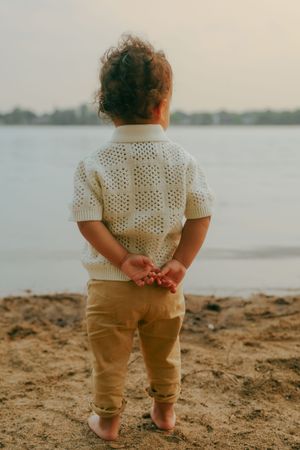 A toddler wearing a white crochet top and khaki pants stands on a sandy beach with hands behind their back at sunset.