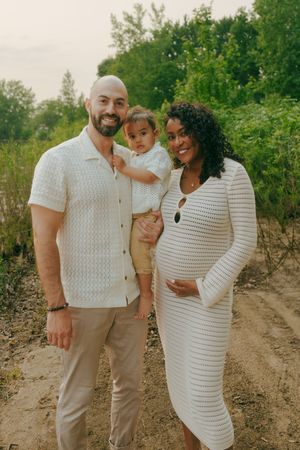 A family shares tender moments together outdoors against a green natural backdrop wearing coordinated white and beige outfits.