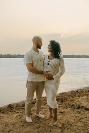 A couple shares an intimate moment at sunset on a sandy beach wearing coordinated white and beige outfits.
