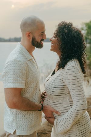 Romantic couple in matching white outfits share intimate moments by the water during sunset photoshoot.