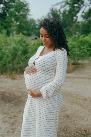 A pregnant woman in a white striped dress cradles her baby bump while standing outdoors in a natural setting.