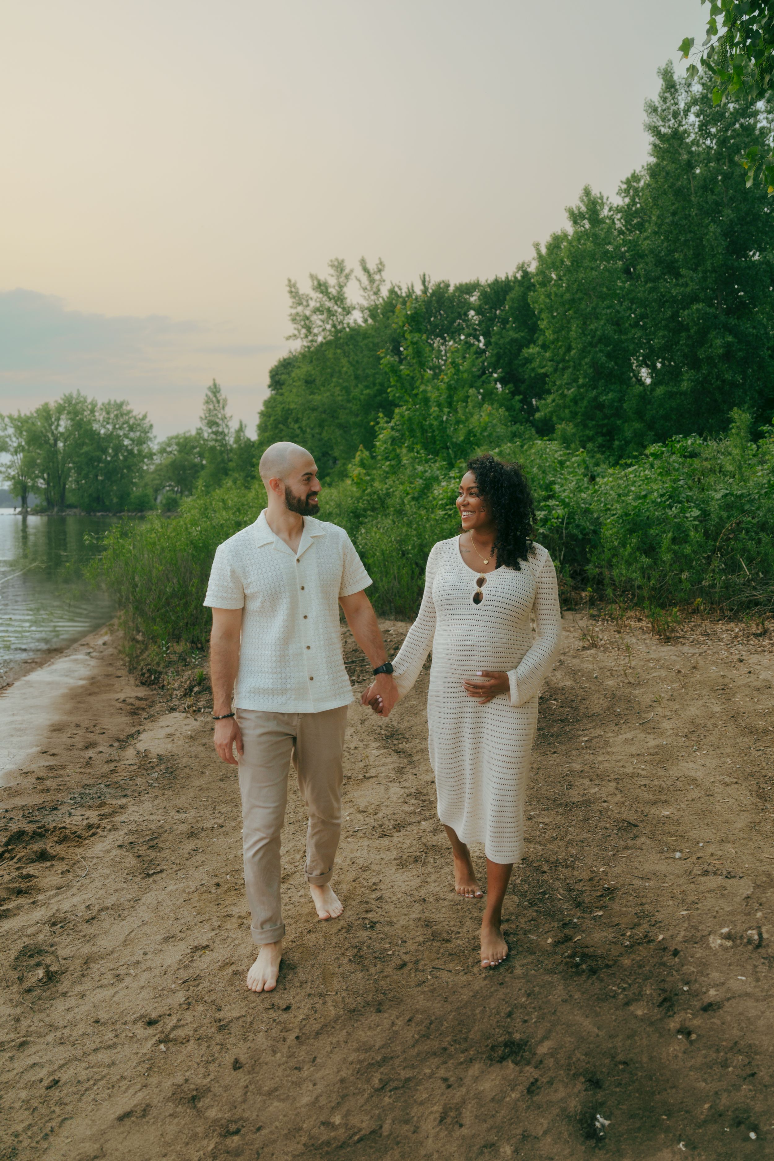 A couple takes a romantic stroll along a tree-lined shore path during golden hour wearing matching light-colored attire.