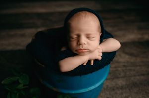 A sleeping newborn in dark clothing rests peacefully on a blue posing pillow against a dark backdrop.