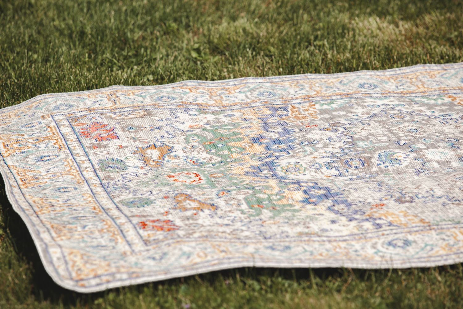 A detailed oriental rug with intricate floral patterns lies on green grass in soft sunlight.