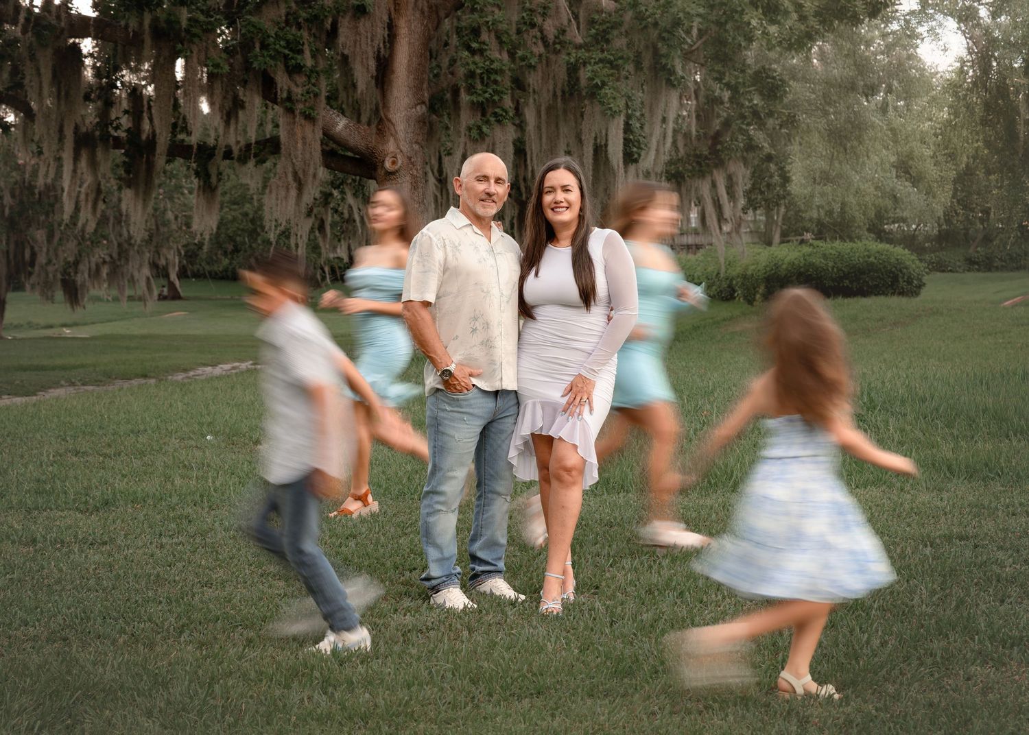 A couple stands on a grassy lawn under moss-draped trees while children play and run around them in motion blur.
