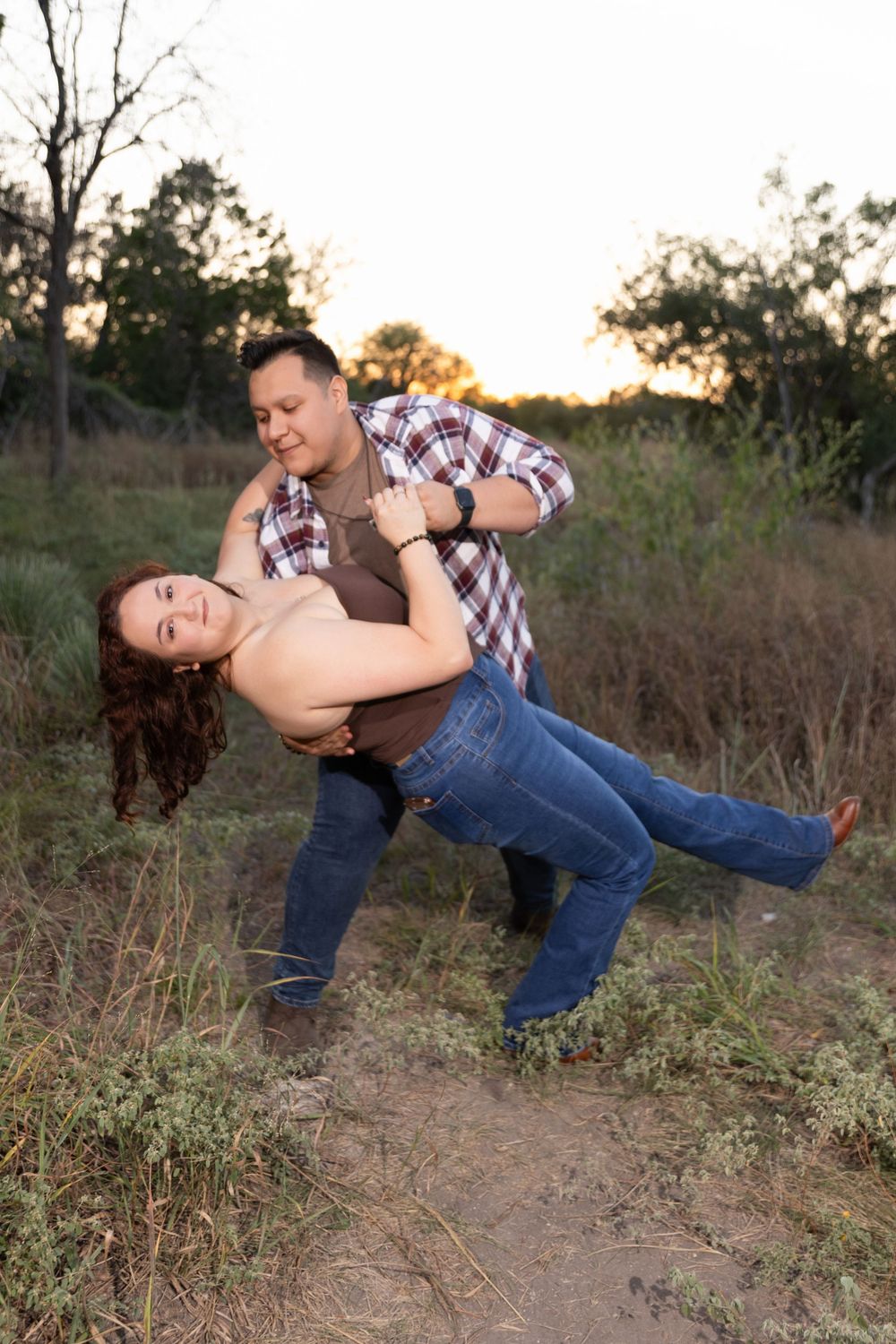 A playful couple dances together in a field at sunset, with one person dipping the other in a romantic pose.
