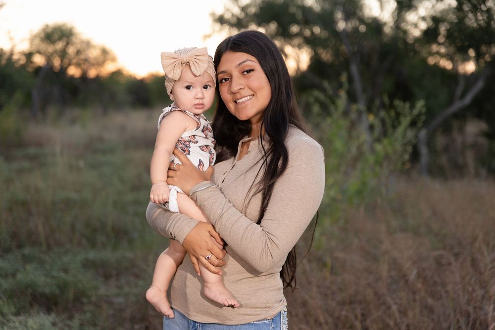 Two people in beige sweaters embrace while standing in a field at sunset.