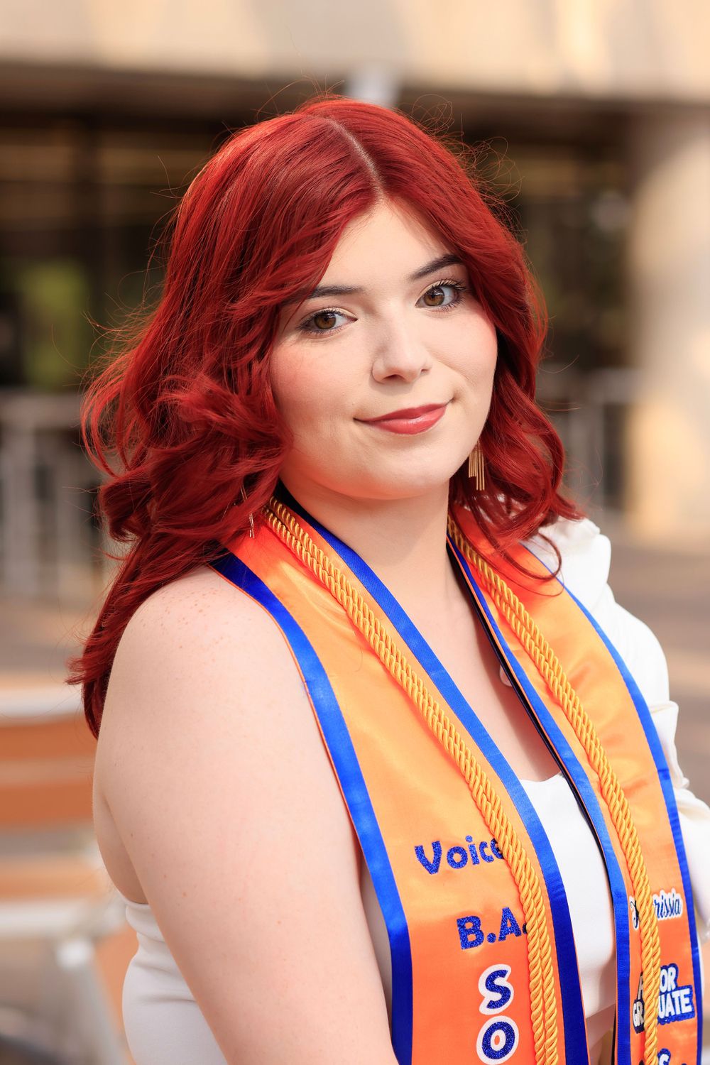 Graduate wearing orange and blue graduation stole smiles at camera.