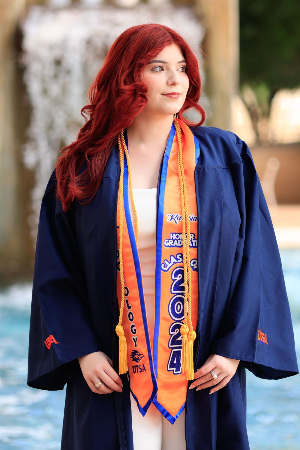 Graduate in navy gown and orange sash poses by fountain.