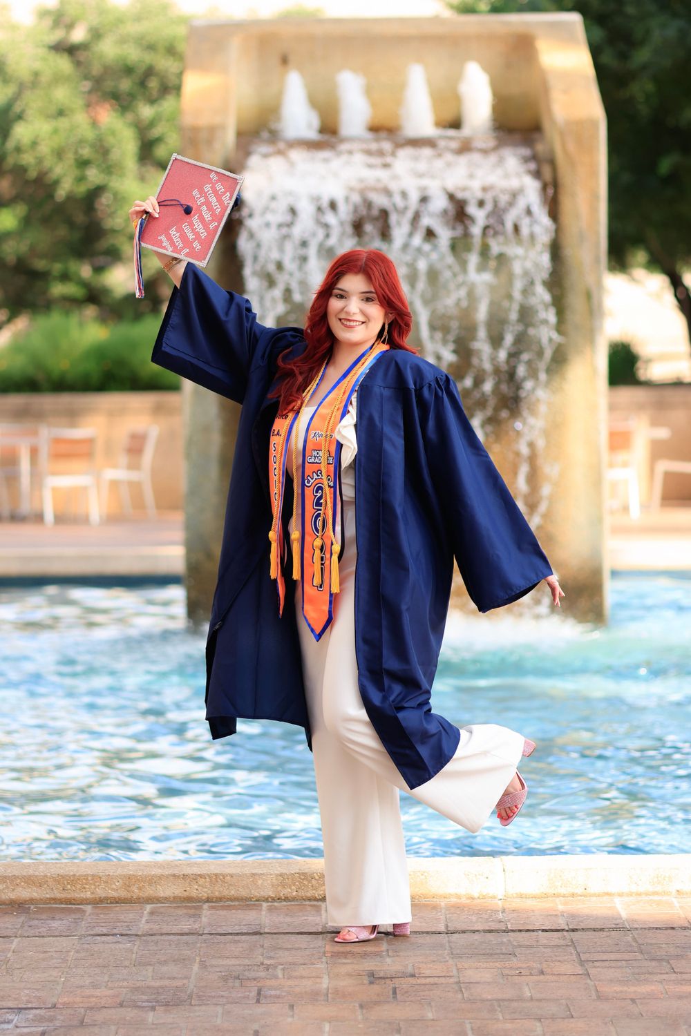 Graduate in blue cap and gown celebrates by fountain with diploma in hand.