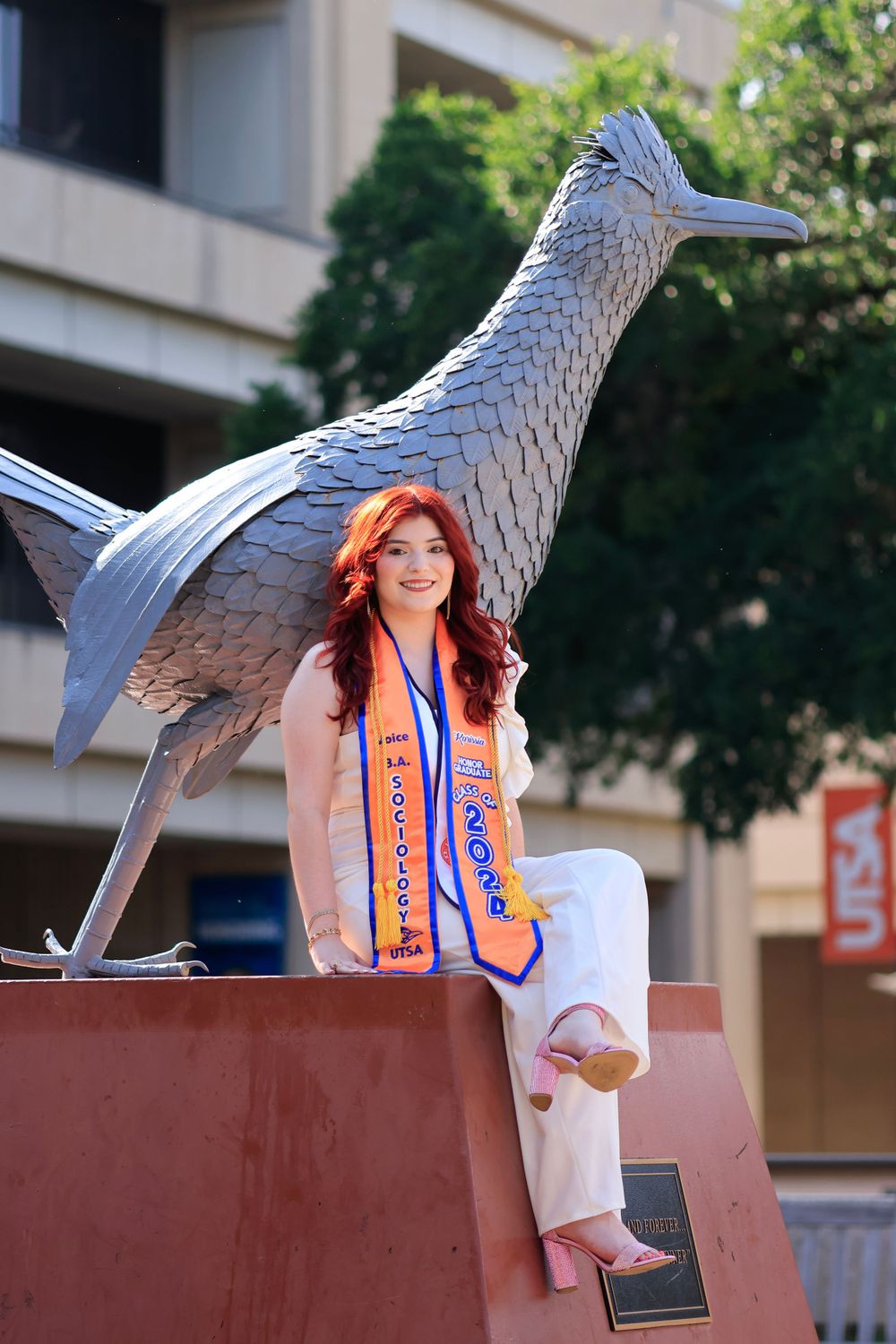 Graduate in white dress poses with large metal bird sculpture on outdoor campus plaza.