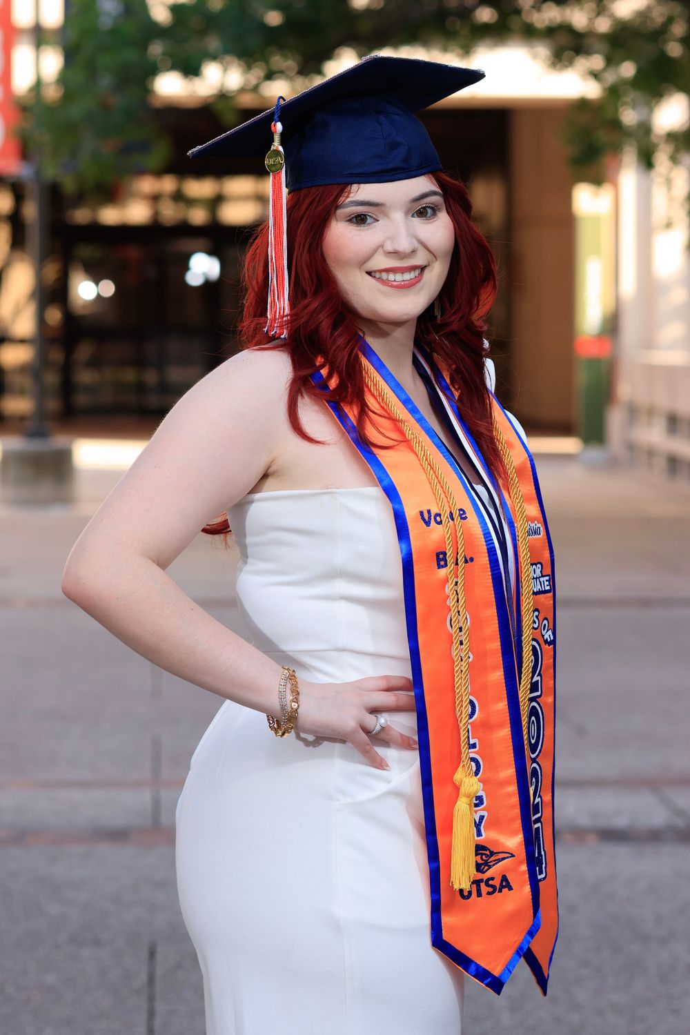 Graduate wearing cap and gown with orange stole poses outdoors during commencement.