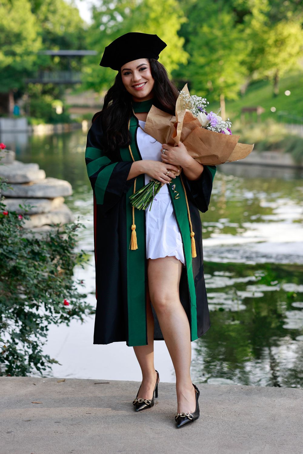 Graduate in green and black academic regalia poses with flower bouquet by scenic pond.