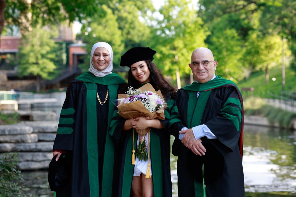 Three people in academic regalia pose together outdoors by stone wall and pond.