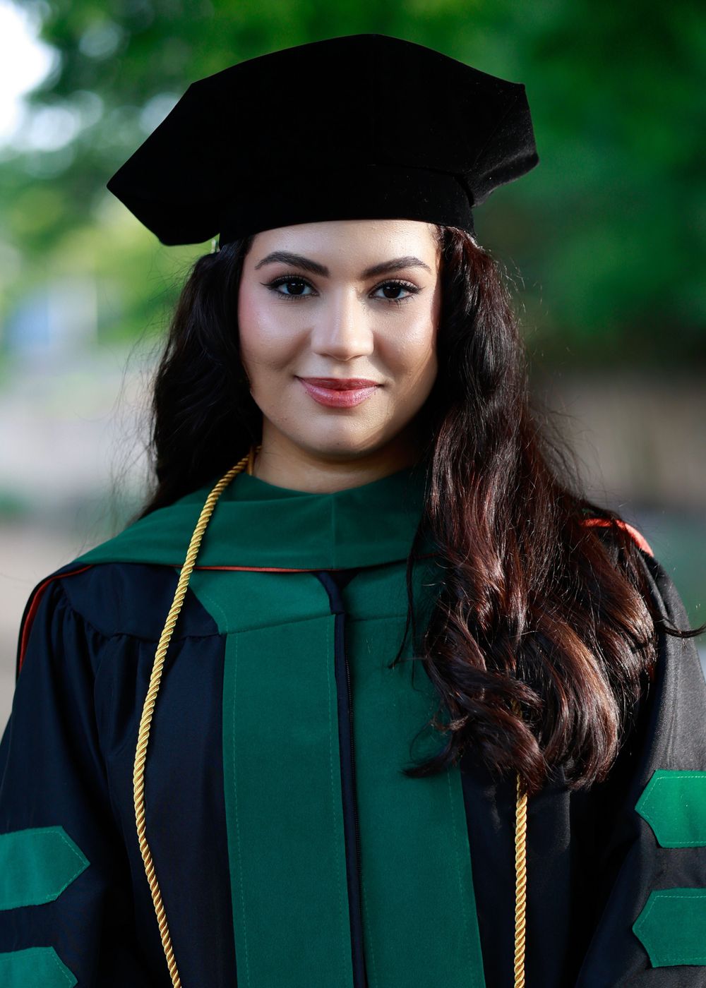 Graduate wearing traditional black cap and green academic regalia poses outdoors.
