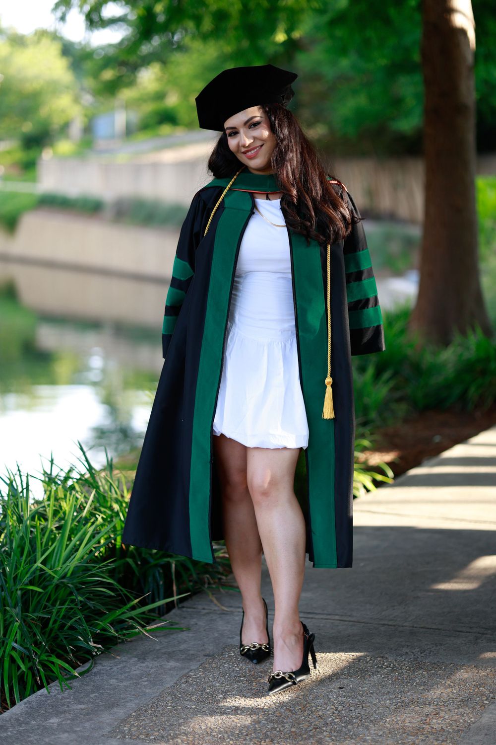 Graduate in black and green academic gown poses on path by water.