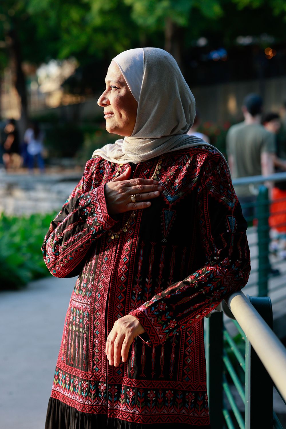 Person in patterned red dress and hijab stands by railing outdoors at sunset.