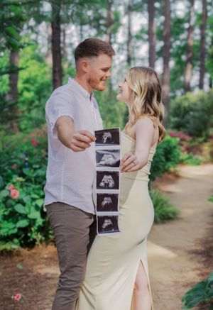 A couple in casual attire shares ultrasound photos on a wooded path.