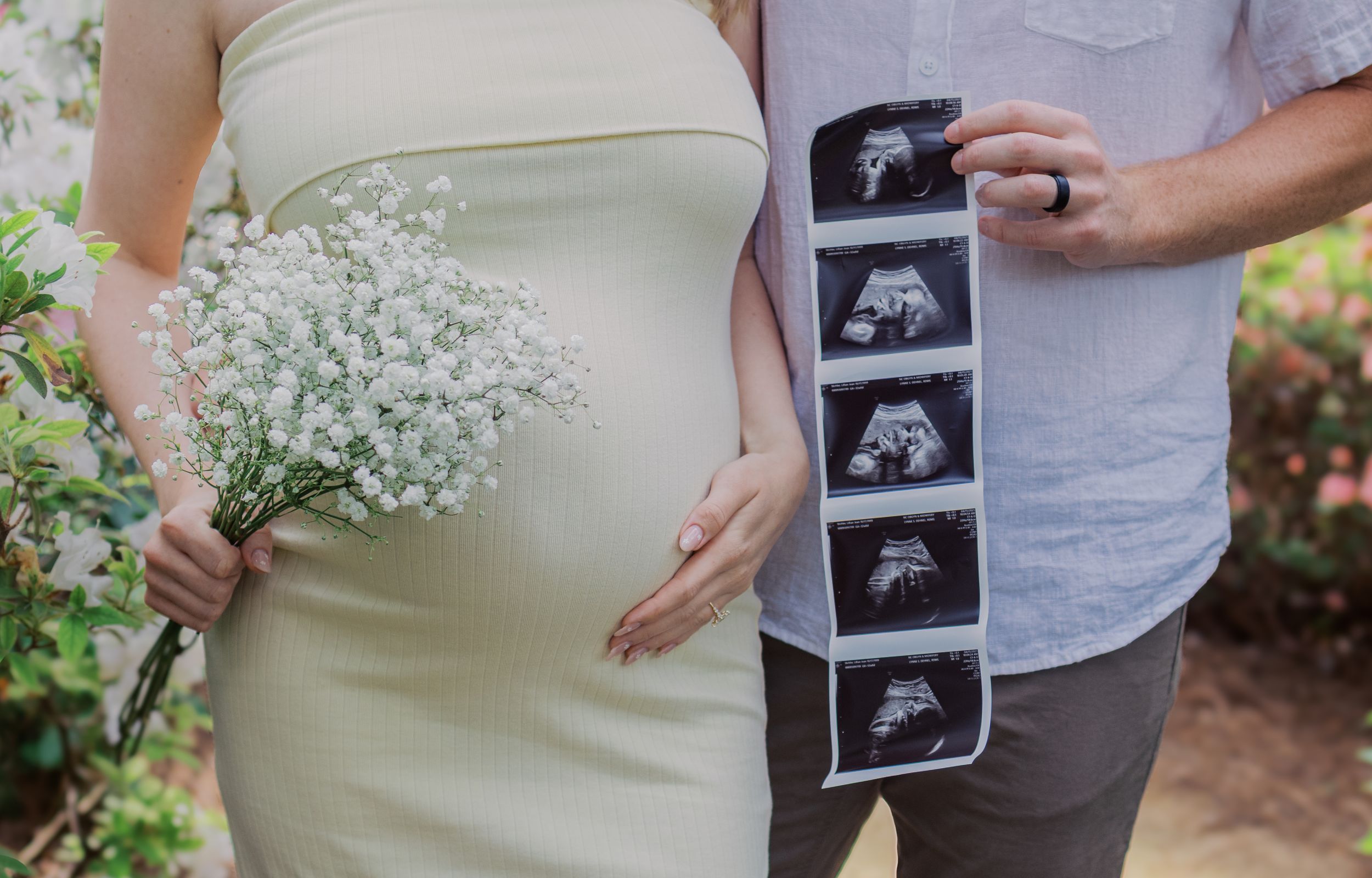 A pregnant woman in a cream dress holds baby's breath flowers while displaying ultrasound photos.
