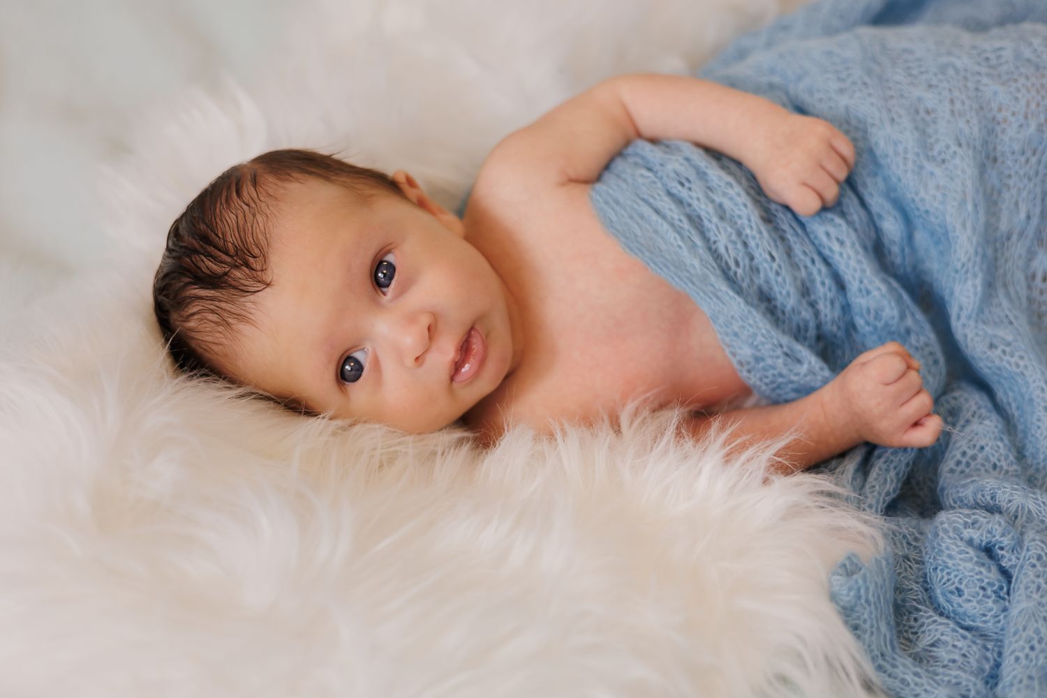 Baby wrapped in soft blue blanket lying on white fur background.