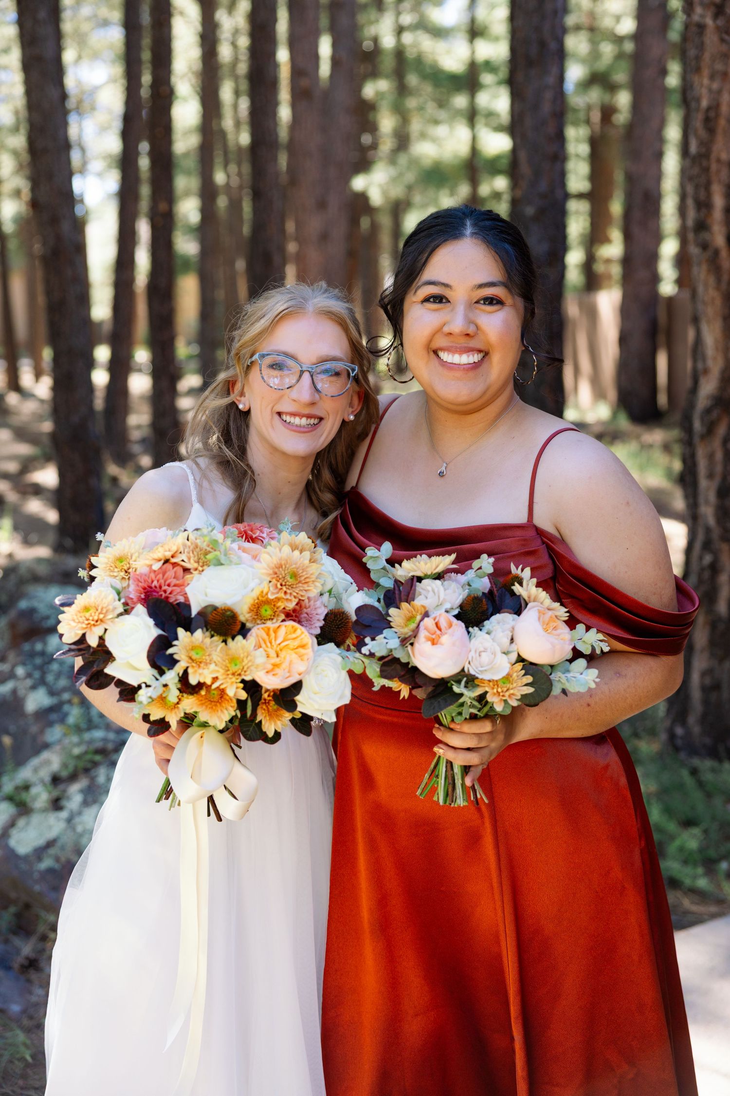 Bridesmaids wearing burgundy gowns and holding orange and white floral bouquets stand together in a forest setting.