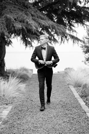 Groom in black tuxedo walking gravel path flanked by ornamental grasses, black and white.