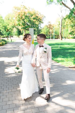 Smiling newlyweds walk hand in hand on park pathway, bride in lace dress, groom in tan suit.