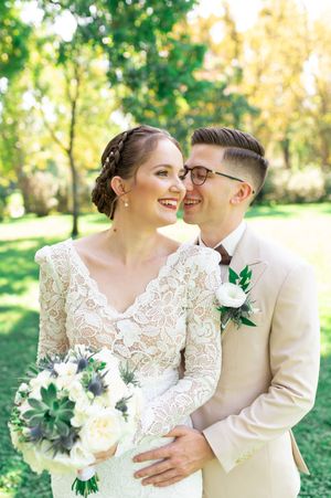 Smiling bride in long-sleeve lace gown holds white bouquet as groom in beige suit whispers in her ear outdoors.