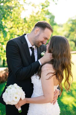 Joyful groom in black suit embracing bride in white gown in sunlit garden setting.