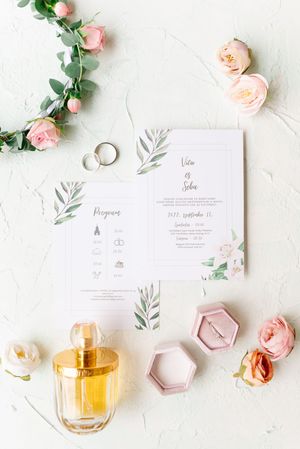 Flat lay of floral wedding stationery, rings, pink roses, perfume bottle, and ring box.