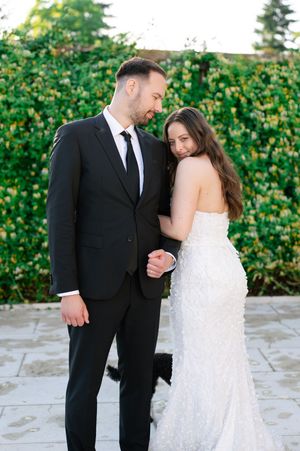 Bride in strapless white gown and groom in black suit sharing intimate moment outdoors.