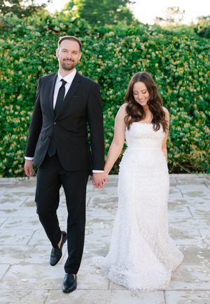 Bride in white lace gown and groom in black suit holding hands walking outdoors.