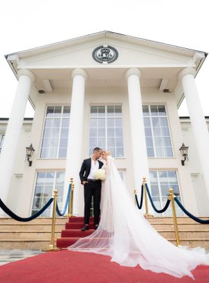 Bride and groom posing on red carpet steps before a grand white columned building with velvet ropes.