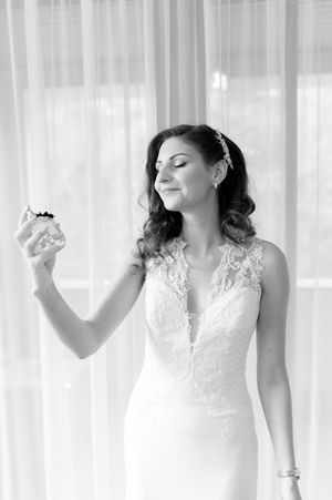 Elegant bride in lace gown holds perfume bottle smiling softly in black and white portrait.