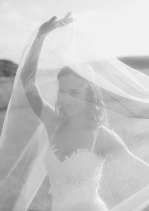 Black and white artistic photo of a smiling bride lifting her flowing veil in an outdoor setting.