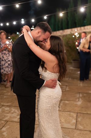 Bride in beaded gown and groom in black tuxedo share passionate kiss under string lights outdoors.