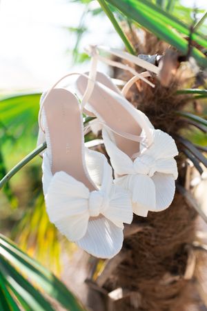 White satin bow bridal heels hanging from a tropical palm tree branch in bright natural sunlight.