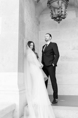 Black and white photo of bride in lace gown holding hands with groom beside stone columns.