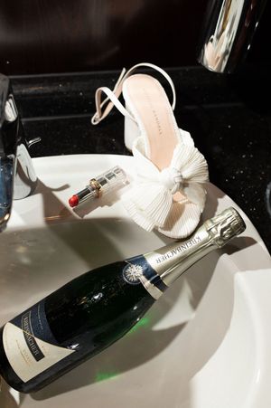 White bow bridal heels and red lipstick rest on sink beside a Taittinger champagne bottle for wedding day prep.