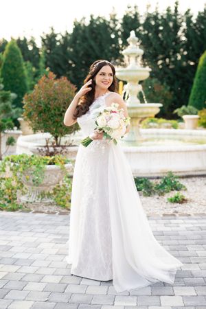 Bride in white lace off-shoulder gown holding floral bouquet by stone fountain in garden.