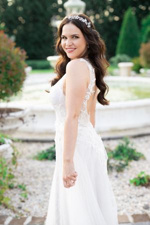 Smiling bride in lace backless wedding gown with crystal hair piece posing gracefully in a sunlit garden.