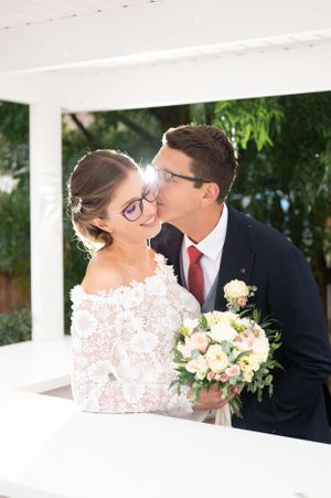 Groom in navy suit kissing smiling bride wearing glasses and lace off-shoulder dress while holding a bouquet.