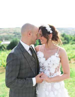 Bride in floral applique gown and groom in plaid suit share an intimate moment at vineyard.