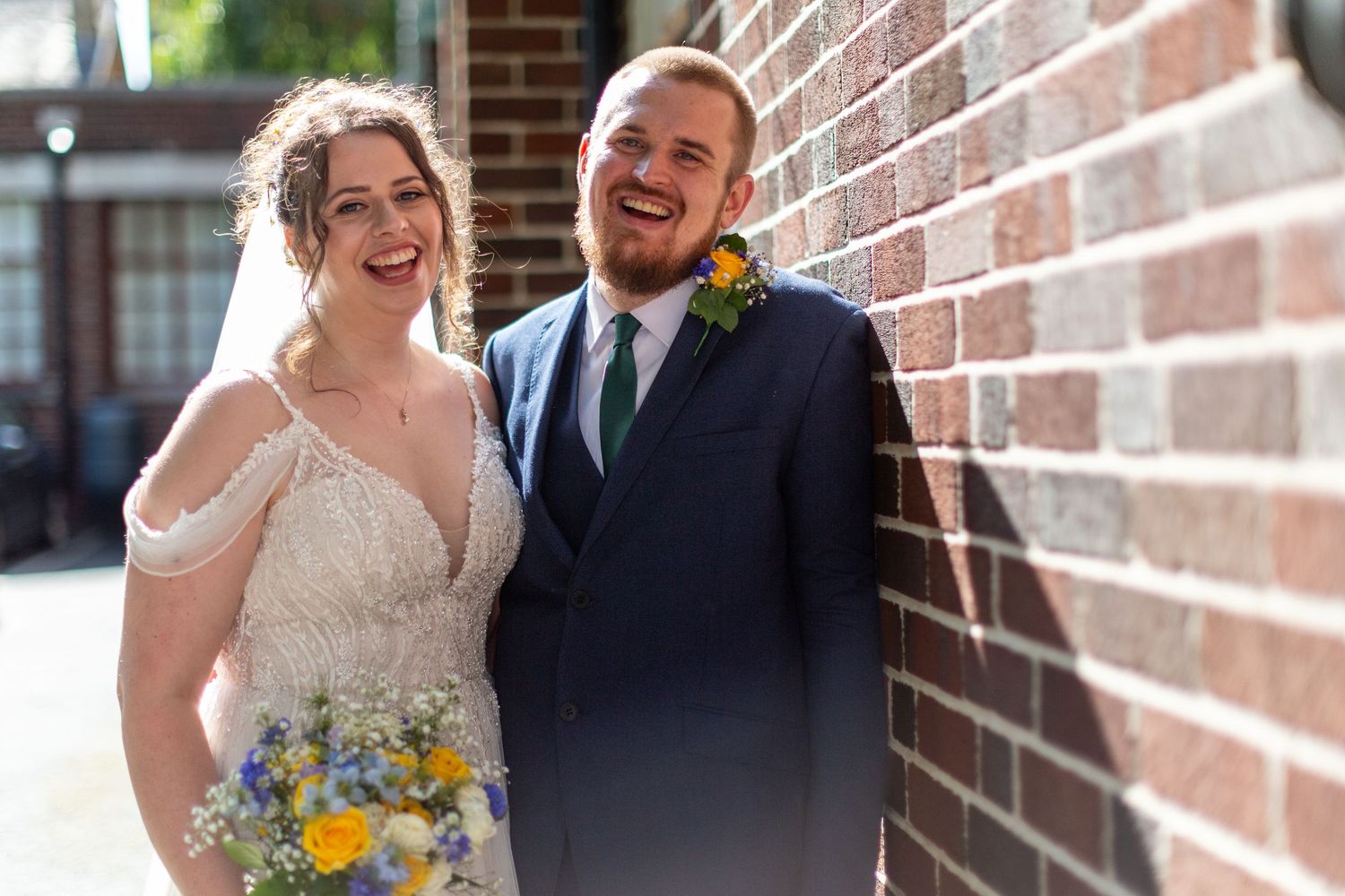 Joyful wedding moment captured against rustic brick wall with yellow floral bouquet.