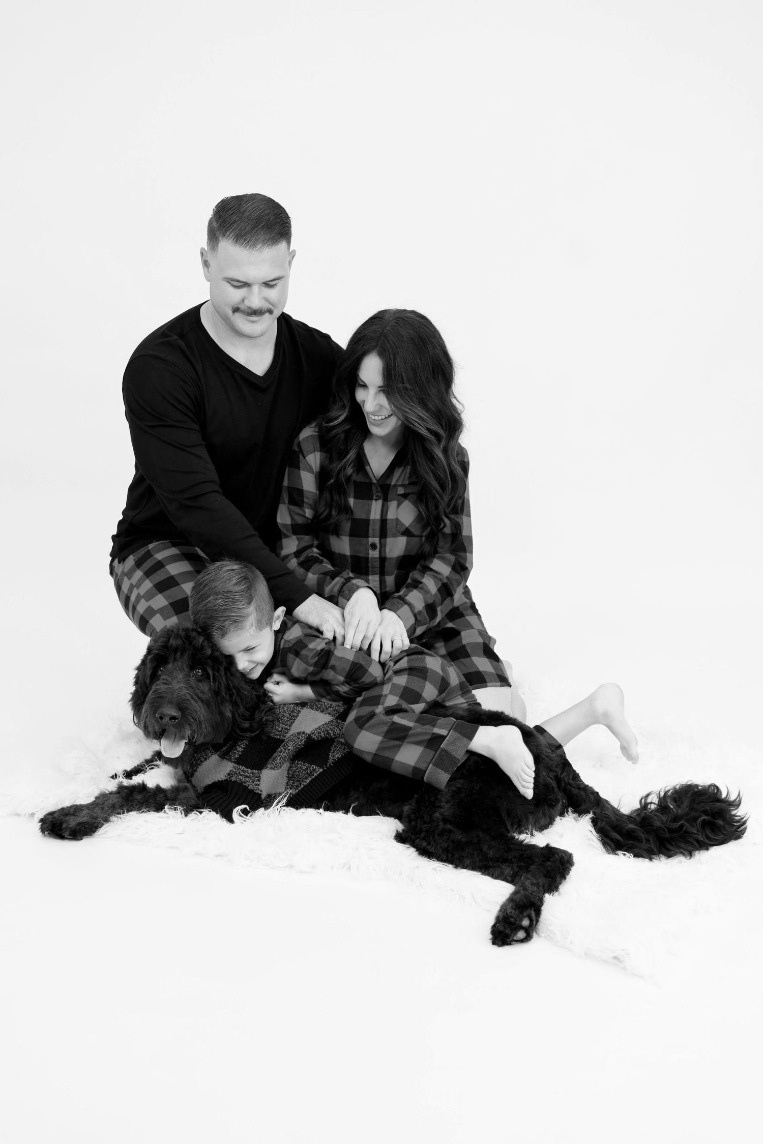 Black and white portrait of people and a dog in matching plaid lounging on white background.