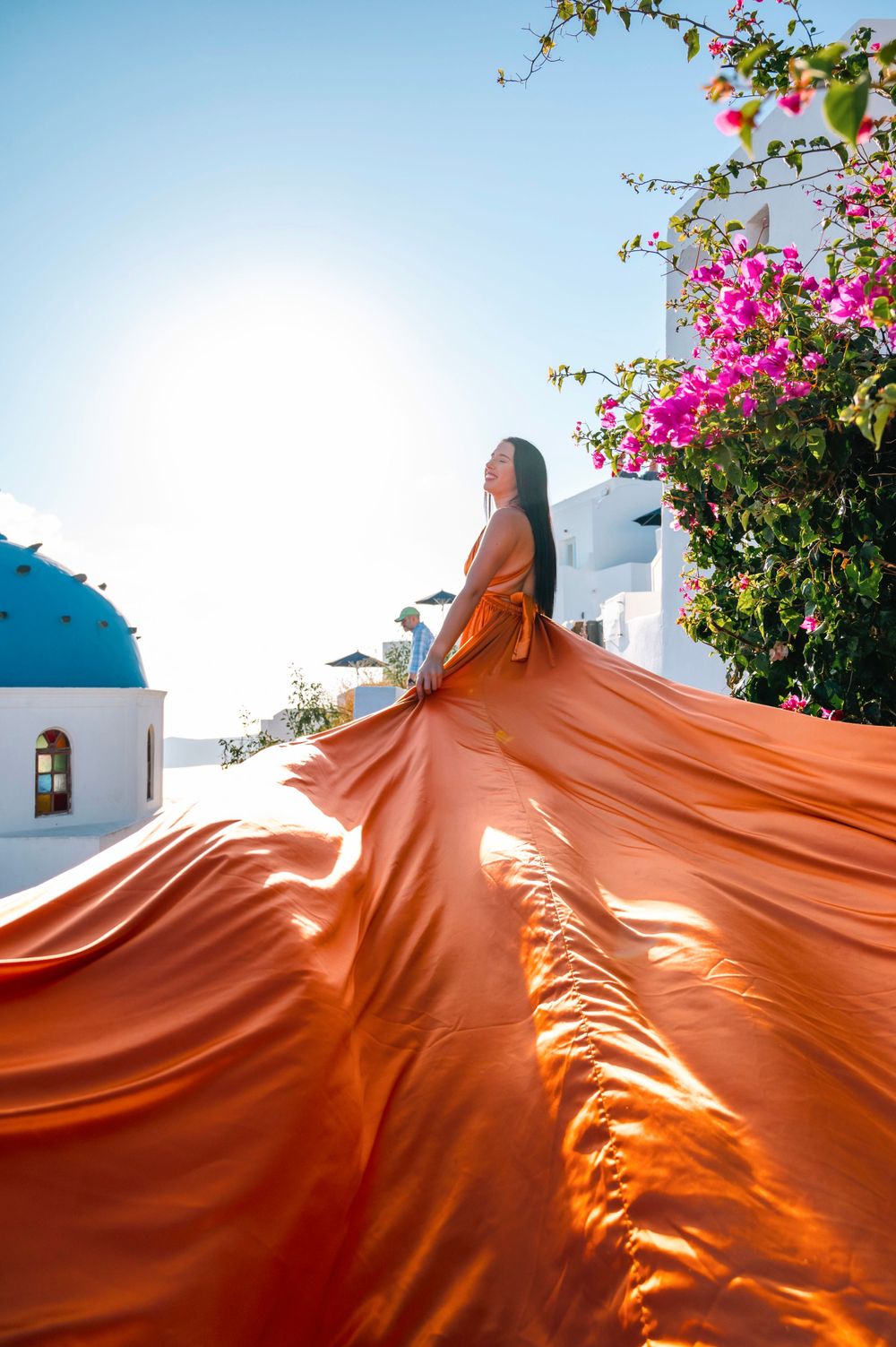 Orange flowing dress contrasts with pink bougainvillea flowers against white Mediterranean architecture.