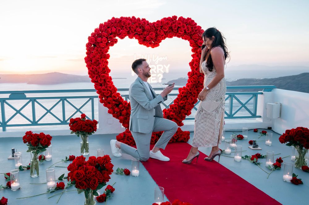 Marriage proposal setup with large red rose heart arch and red carpet overlooking ocean at sunset.