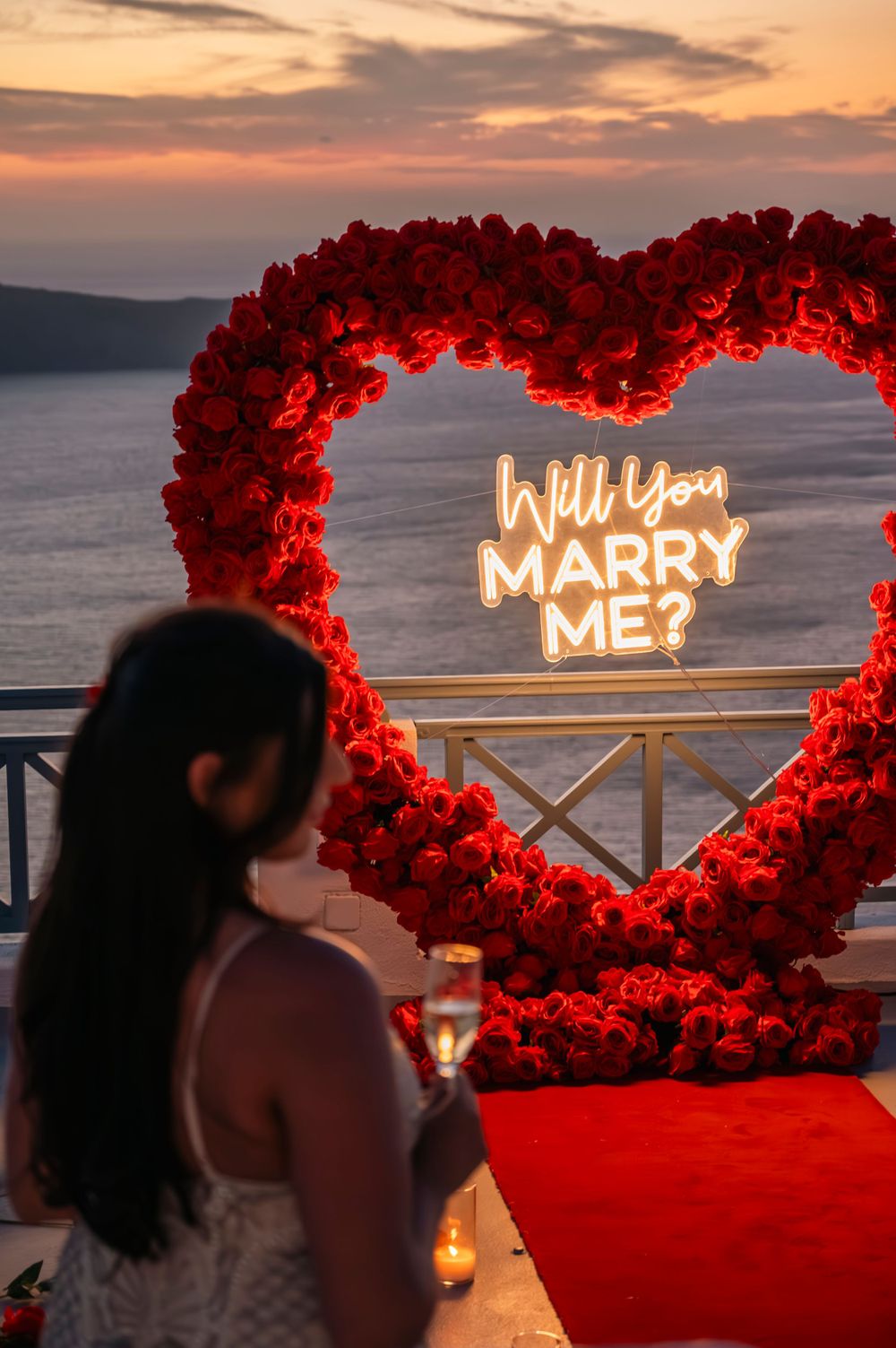 Red heart-shaped arch with illuminated 'Will you marry me?' sign at sunset.