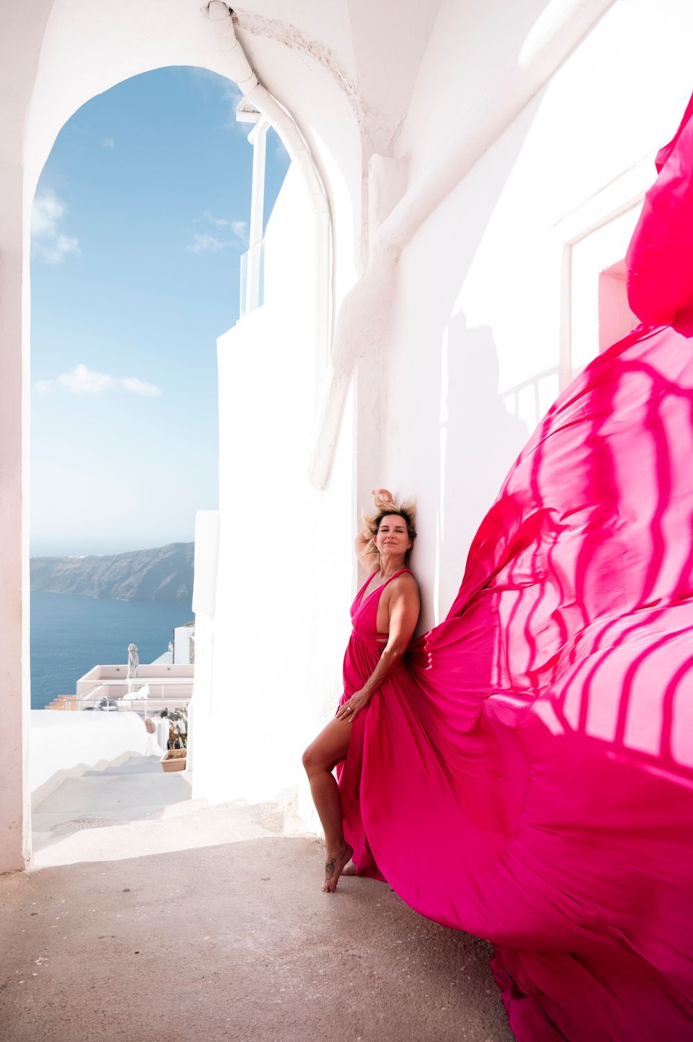 Flowing hot pink dress billows dramatically in white Mediterranean archway overlooking sea.